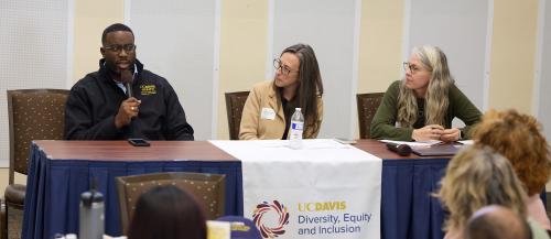 African American man sits at table and speaks into microphone to crowd seated at round tables. Two women, who are seated next to him, look at him as he speaks.