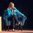 Cynthia Miller-Idriss speaks at the Mondavi Center on Feb. 5. She is seated in a blue chair on stage and is wearing a bright teal coat and black pants and is speaking into a microphone.